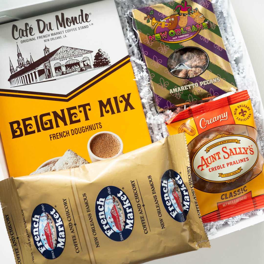 Photograph of a New Orleans themed gift basket featuring a yellow Café Du Monde beignet mix box showing the St. Louis Cathedral landmark, a gold-foiled French Market coffee pouch, and purple amaretto pecan packaging nestled in white shredded paper