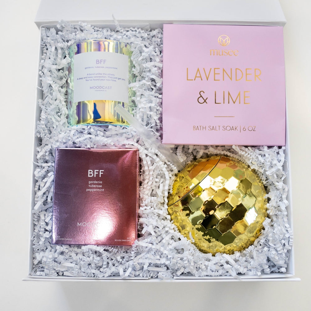 Digital photograph of a curated luxury spa gift set featuring an iridescent Moodcast BFF candle, a pink Musce Lavender and Lime bath soak box, and a reflective golden disco ball resting on white crinkle paper shred