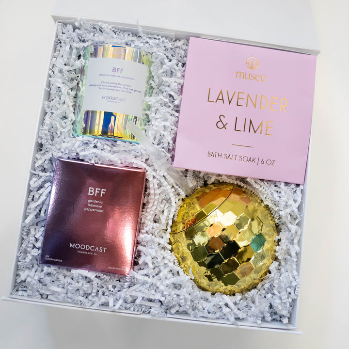 Digital photograph of a curated gift box featuring a pink Musee Lavender &amp; Lime bath soak, an iridescent Moodcast BFF candle jar, and a shiny gold disco ball tumbler nestled in white crinkle paper