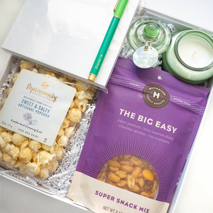 Photograph of a curated gift box featuring a purple bag of Hammond’s snack mix and Popinsanity popcorn nestled next to a light green Cactus Flower candle and a vibrant teal pen on white crinkle paper filler