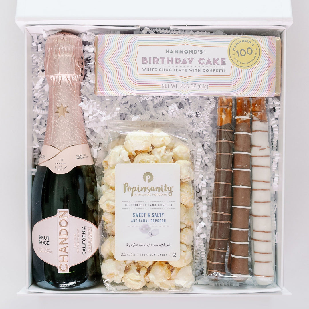 Product photograph of a celebratory gift box featuring a Chandon Brut Rosé bottle with pink foil, a Hammond’s Birthday Cake chocolate bar in a pastel rainbow wrapper, Popinsanity popcorn, and three chocolate-dipped pretzels on white crinkle paper filler