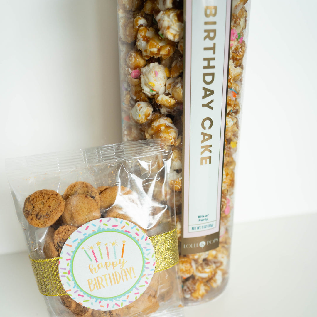 Photograph from Lolli &amp; Pops of a tall cylinder of gourmet Birthday Cake popcorn and a small bag of mini chocolate chip cookies on a white surface, featuring a gold glitter ribbon, a colorful sprinkle-themed Happy Birthday sticker, and popcorn clusters drizzled with white icing and rainbow sprinkles