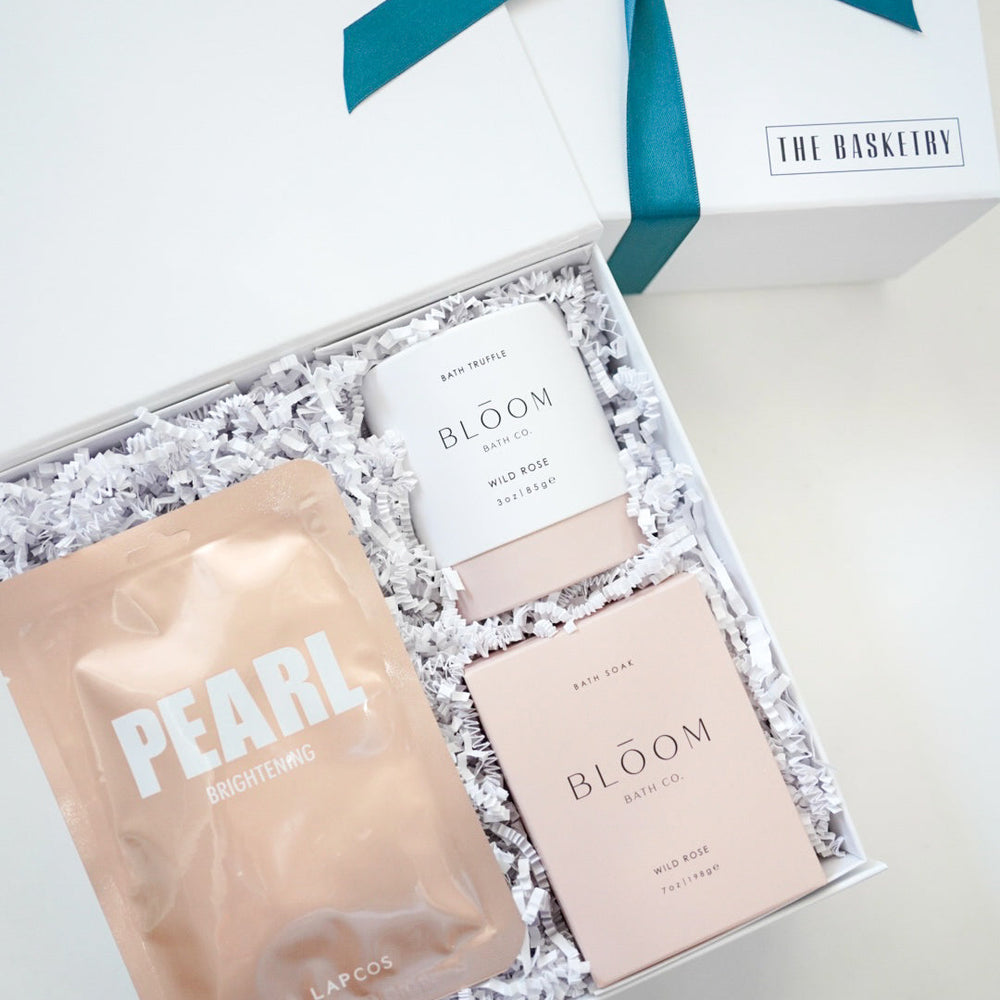 Digital photograph by The Basketry of a white spa gift box containing a rose gold Lapcos Pearl mask and pink Bloom Bath Co. wild rose bath products nestled in white crinkle paper with a teal ribbon on the lid