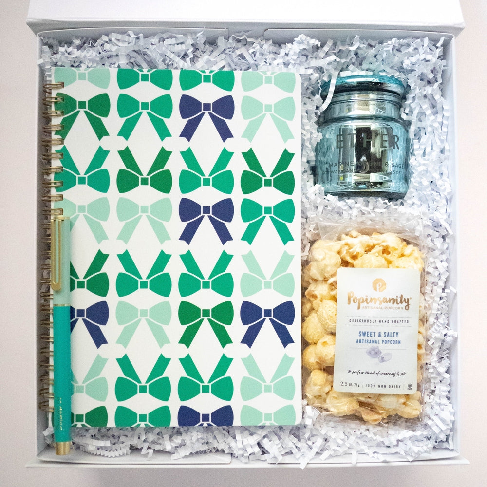 Photograph of a curated gift box featuring a notebook with a repeating teal and navy bow pattern, a matching teal pen with gold accents, an Ether light blue glass candle, and a bag of Popinsanity Sweet & Salty popcorn nestled in white crinkle paper