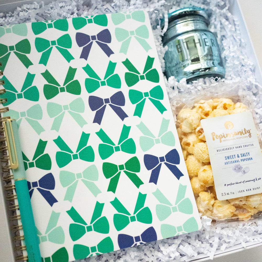 Photograph of a curated gift box featuring a spiral notebook with a mint and navy bow pattern, a matching teal pen, a blue glass candle, and Popinsanity popcorn nestled in white crinkle paper