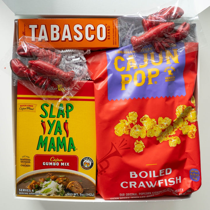 Photograph of a Louisiana-themed Cajun food gift box featuring iconic Tabasco, Slap Ya Mama, and Boiled Crawfish popcorn brands alongside bright red plastic crawfish and vibrant yellow packaging