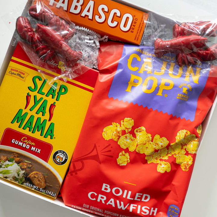 Photograph of a Louisiana-themed food gift basket featuring Slap Ya Mama gumbo mix, Tabasco, and Cajun Pop popcorn alongside red crawfish-shaped candies and white shredded paper filler