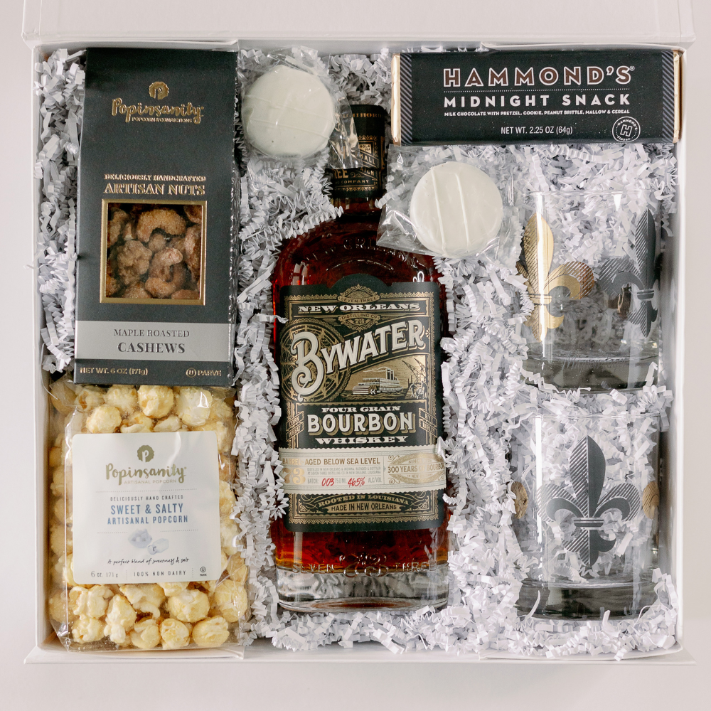Studio photograph of a New Orleans-themed gourmet gift box featuring a bottle of Bywater Four Grain Bourbon with a river steamboat on the label, two whiskey glasses with gold fleur-de-lis motifs, and artisan snacks nestled in white crinkle-cut paper