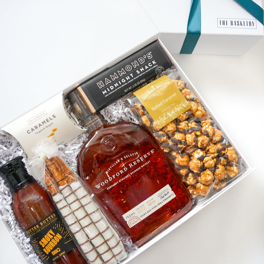 Gift set with a bottle of whiskey, snacks, and a bottle of hot sauce on a white surface.