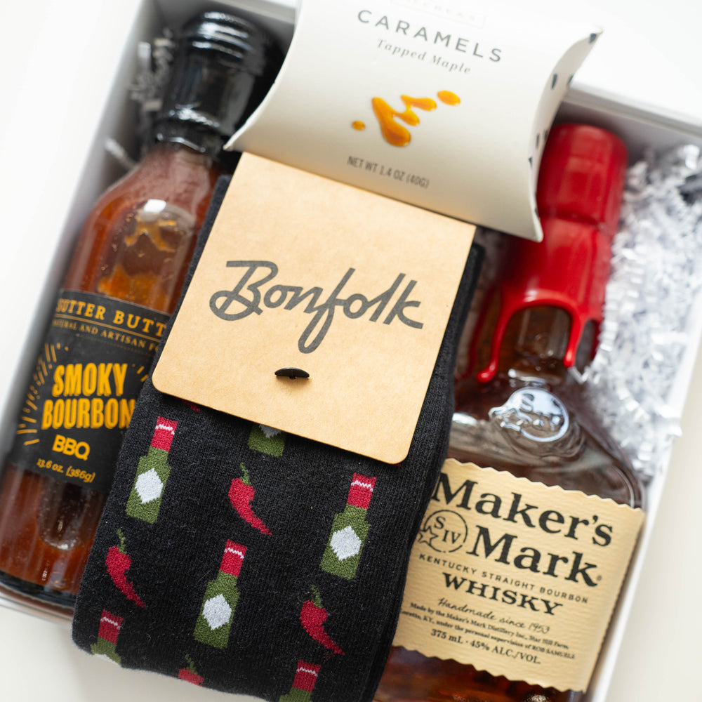 Commercial photograph of a curated bourbon gift box featuring a bottle of Maker's Mark Whisky with its signature red wax seal, Sutter Buttes Smoky Bourbon BBQ sauce, and black Bonfolk socks with a colorful red chili pepper and hot sauce bottle pattern