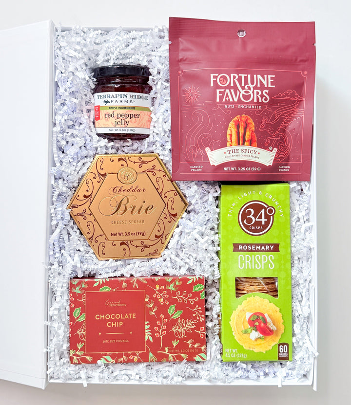 An open white gift box filled with shredded white paper and containing assorted gourmet snacks and treats, including a red bag labeled Fortune Favors, a small jar, a hexagonal box labeled Brie, a red chocolate bar, and a green bag of 345 Crisps, photographed on a light neutral background.