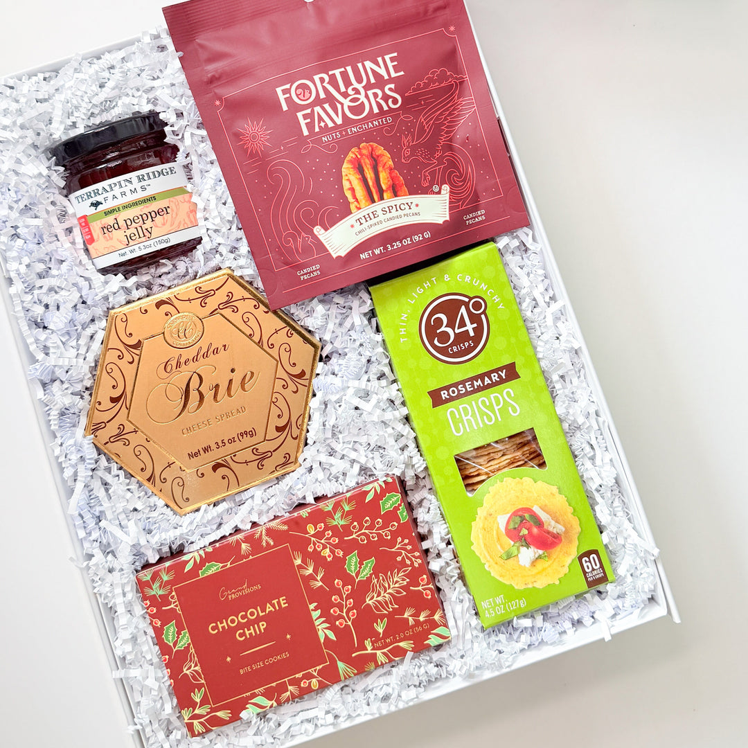 A curated gift box assortment from The Basketwork, featuring a white box with a red ribbon, a red Fortune Favors bag, a green 34% Crikets snack box, a gold Brie cheese card, and a chocolate chip bar, arranged in white crinkle paper.