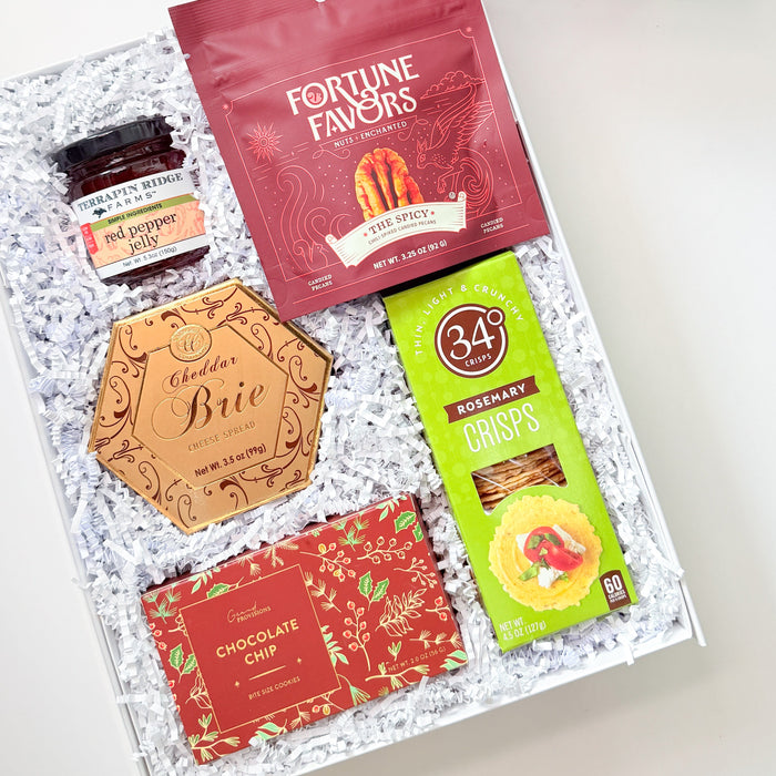 A curated gift box assortment from The Basketwork, featuring a white box with a red ribbon, a red Fortune Favors bag, a green 34% Crikets snack box, a gold Brie cheese card, and a chocolate chip bar, arranged in white crinkle paper.