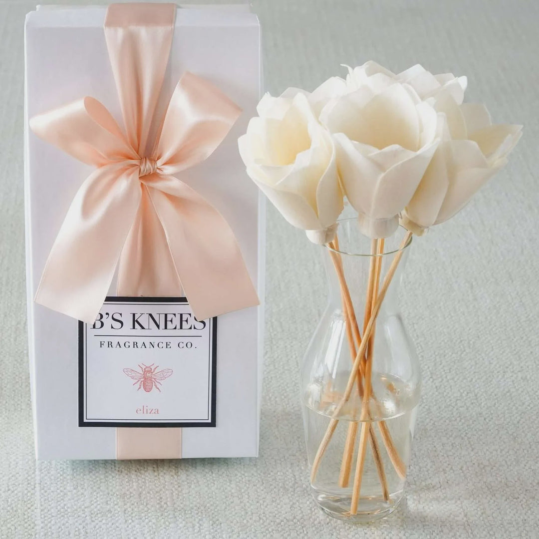 Product photography for B's Knees Fragrance Co. showcasing the Eliza scent gift box with a large peach satin bow and a clear glass vase holding white sola wood flower reed diffusers on a neutral textured background