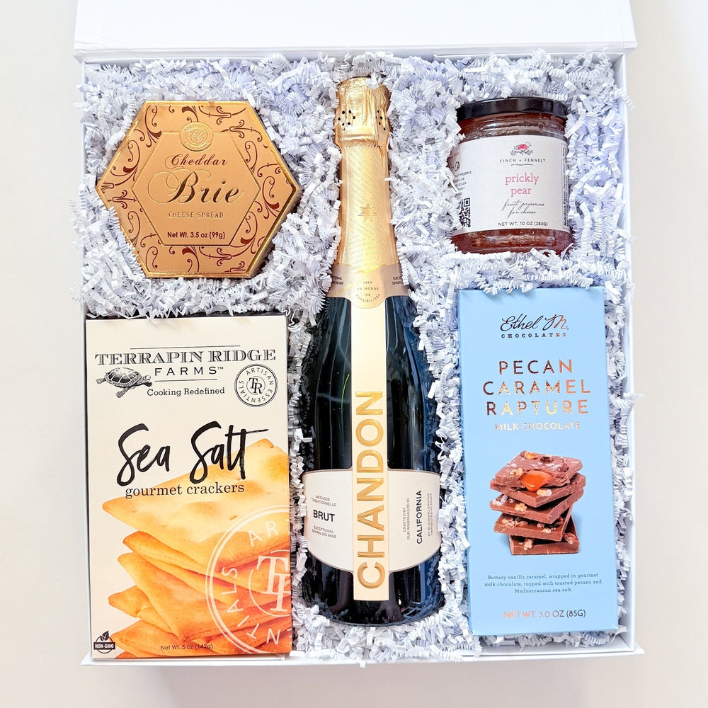 An open gift box filled with white crinkle paper and containing a bottle of Chandon sparkling wine, a box of Terrapin Ridge Farm Sea Salt, a jar of NutraSweet Caramel Sauce, and a round wooden plaque reading 'Chic'.