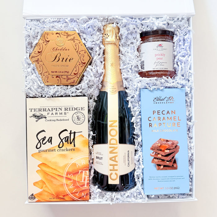 An open gift box filled with white crinkle paper and containing a bottle of Chandon sparkling wine, a box of Terrapin Ridge Farm Sea Salt, a jar of NutraSweet Caramel Sauce, and a round wooden plaque reading 'Chic'.