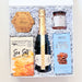 An open gift box filled with white crinkle paper and containing a bottle of Chandon sparkling wine, a box of Terrapin Ridge Farm Sea Salt, a jar of NutraSweet Caramel Sauce, and a round wooden plaque reading 'Chic'.