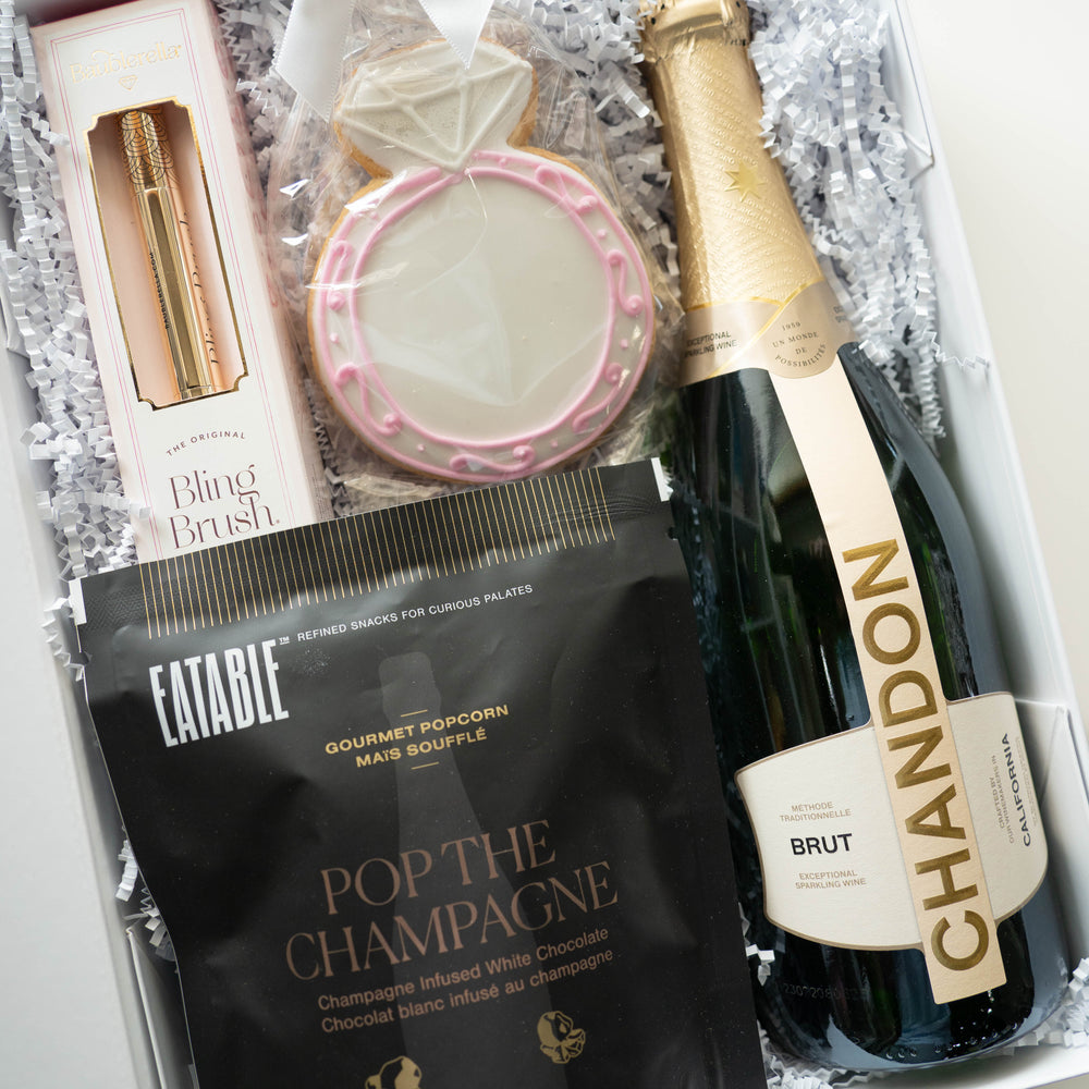 Digital photograph of a curated bridal gift box containing a bottle of Chandon Brut, a Baublerella Bling Brush, and a large diamond ring-shaped sugar cookie with pink icing swirls nestled in white shredded paper