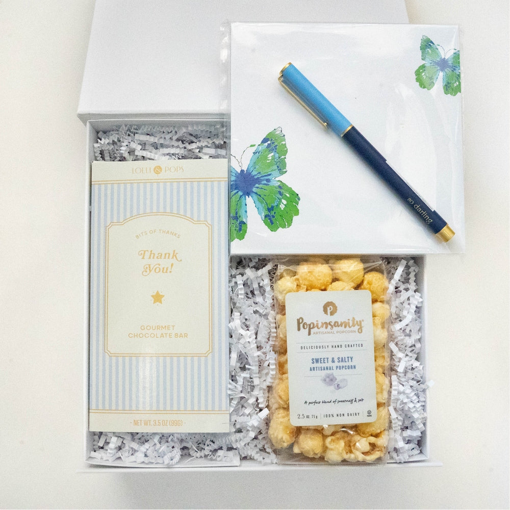 Photograph of a curated gift box featuring a Lolli &amp; Pops gourmet chocolate bar, Popinsanity sweet and salty popcorn, a notepad with vibrant blue and green butterfly illustrations, and a gold-accented blue pen resting on white crinkle paper bedding