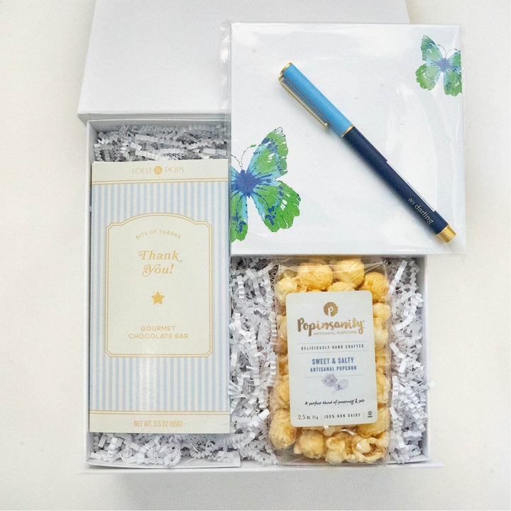 Photograph of a curated gift box featuring a Lolli &amp; Pops gourmet chocolate bar, Popinsanity sweet and salty popcorn, a notepad with vibrant blue and green butterfly illustrations, and a gold-accented blue pen resting on white crinkle paper bedding