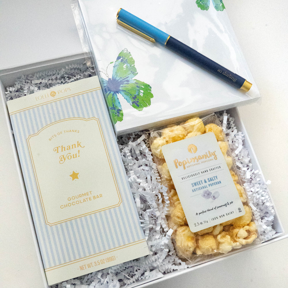 Photograph of a Lolli & Pops and Popinsanity thank you gift set featuring a light blue striped chocolate bar, artisanal sweet and salty popcorn, and a watercolor butterfly notepad with a navy blue so darling pen nestled in white crinkle paper