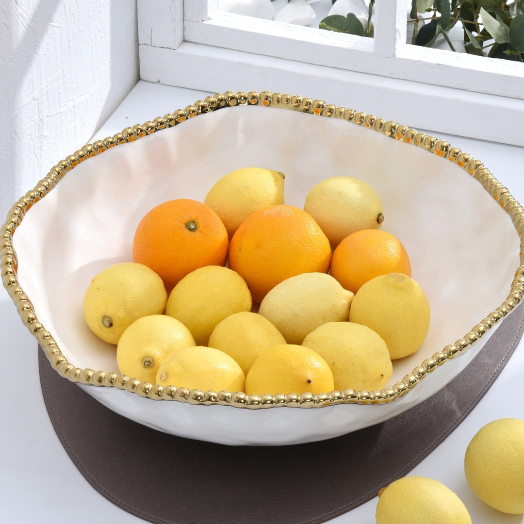 Photograph of a wavy white ceramic bowl featuring a decorative gold beaded rim filled with vibrant yellow lemons and oranges sitting on a grey mat near a white window frame