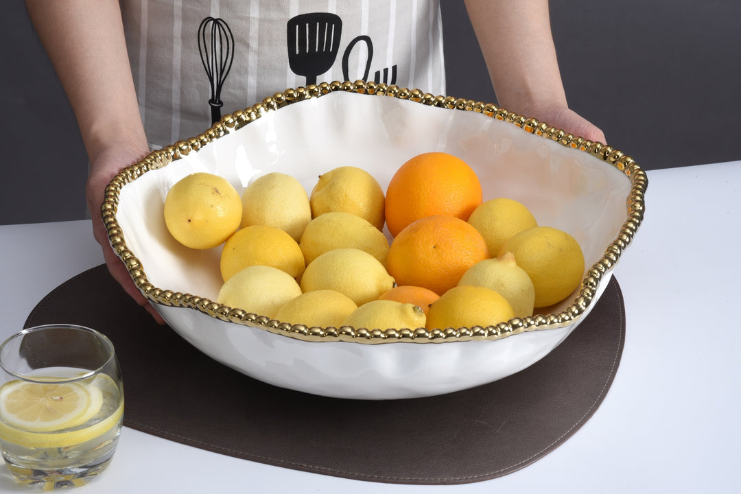 Photograph of a wavy white ceramic fruit bowl with a gold beaded rim filled with lemons and oranges held by a person in a striped apron next to a glass of lemon water
