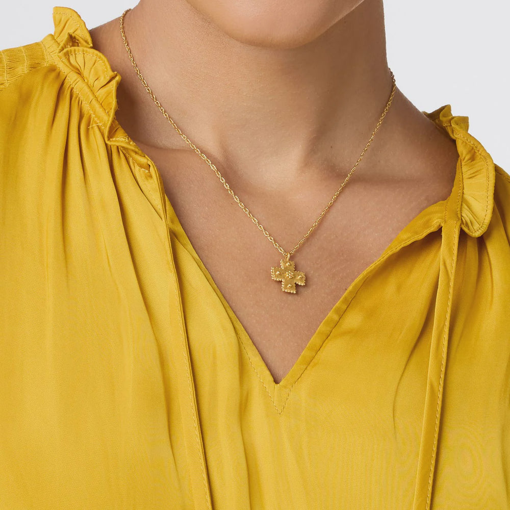 Gold jewelry photograph featuring a textured cross pendant worn on a woman's neck against a mustard yellow ruffled blouse with intricate gold granulation and a delicate rope-link chain