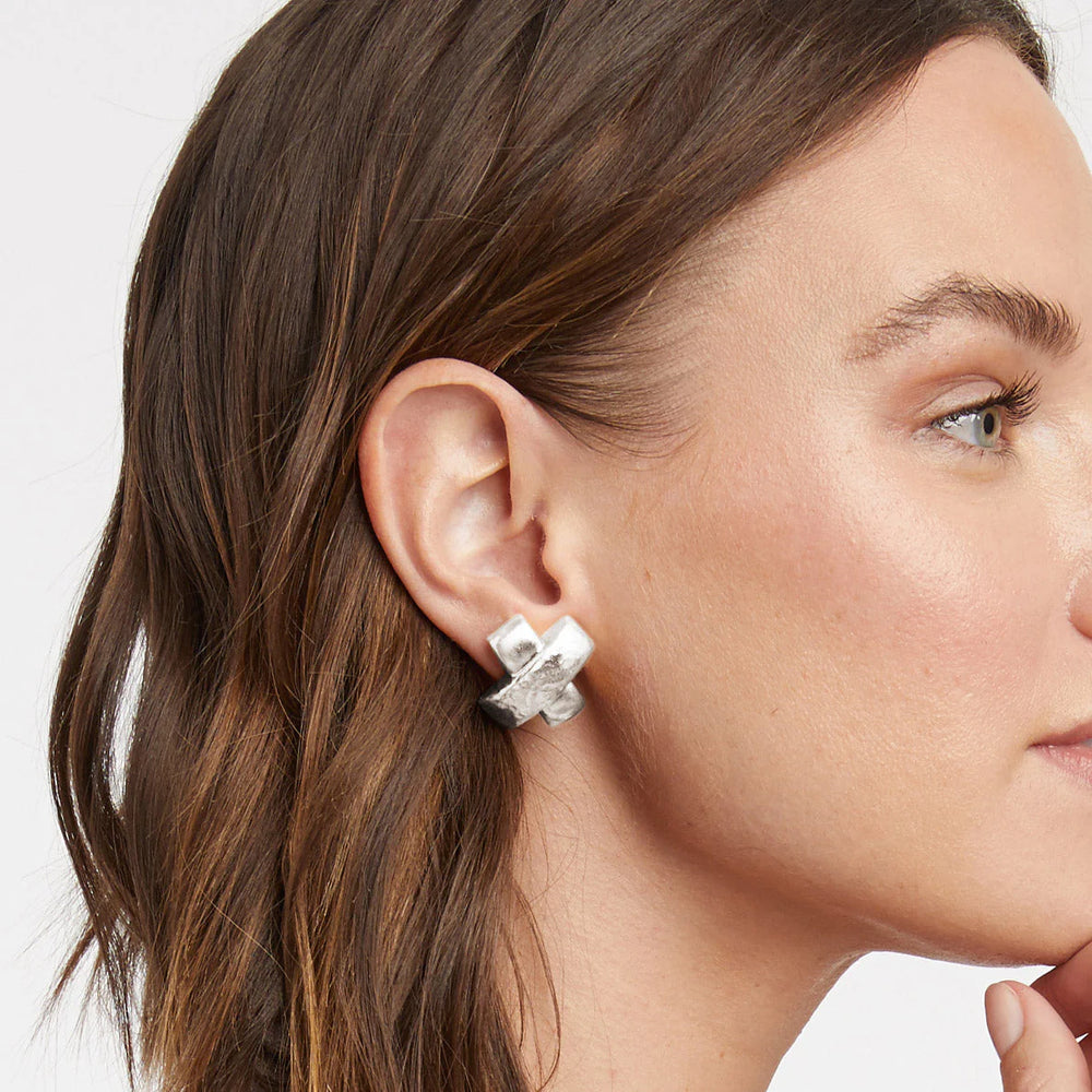 Silver hammered metal earring in an X-shape worn on a woman's earlobe featuring a chunky three-dimensional design and a textured metallic finish