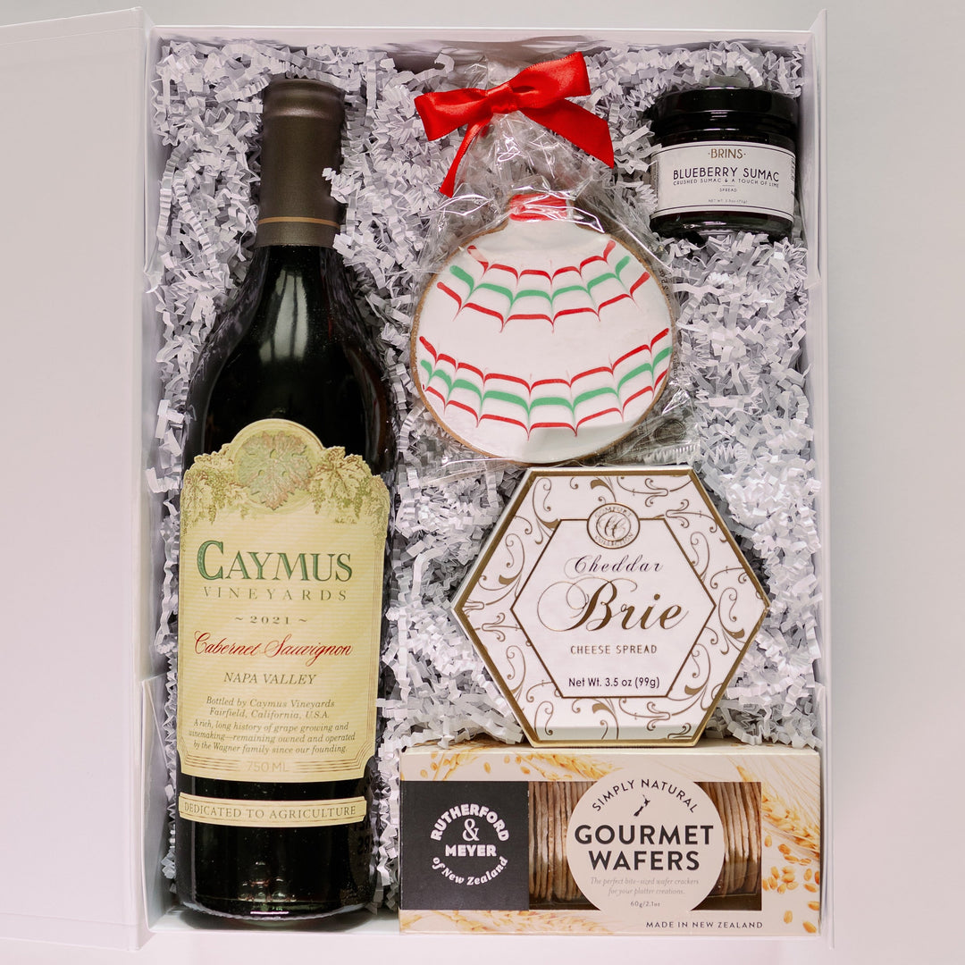 Photograph of a luxury gourmet gift box featuring a bottle of Caymus Vineyards Cabernet Sauvignon, Rutherford & Meyer wafers, and a festive red and green iced holiday cookie tied with a bright red ribbon on white crinkle paper