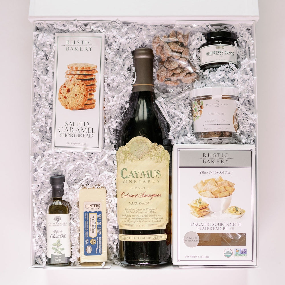 Photograph of a luxury gourmet gift hamper featuring a gold-labeled bottle of Caymus Vineyards Cabernet Sauvignon from Napa Valley surrounded by boxes of Rustic Bakery shortbread and small jars of artisanal preserves nestled in white shredded paper