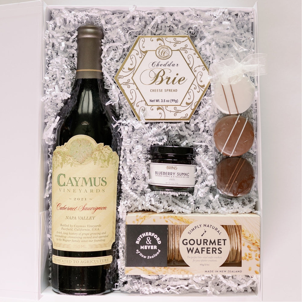 A photograph of a luxury gourmet gift box featuring a bottle of Caymus Vineyards Cabernet Sauvignon from Napa Valley alongside a gold-trimmed hexagonal cheese spread and three chocolate-covered sandwich cookies with white icing stripes nestled in white crinkle paper