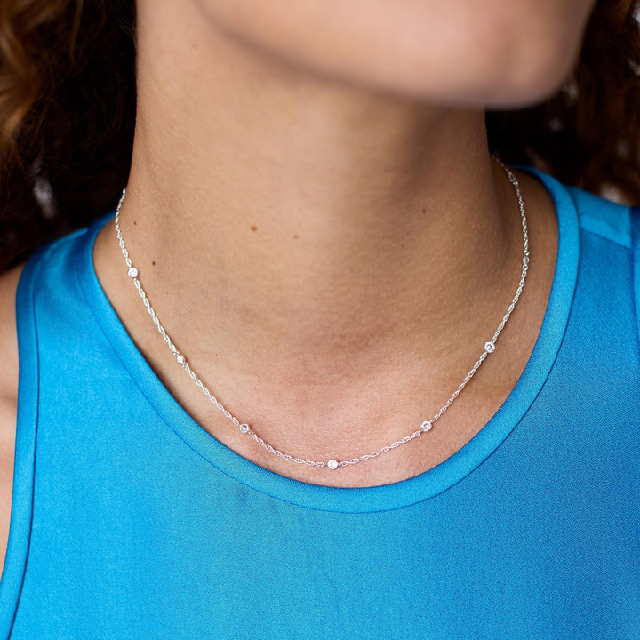 Photograph of a delicate silver station necklace featuring shimmering bezel-set round diamonds on a twisted rope chain worn by a woman in a bright turquoise blue satin top