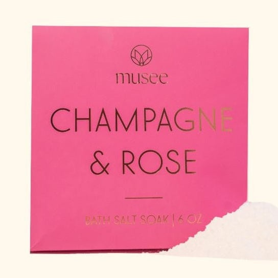 Product photograph by Musee of a Champagne & Rose bath salt soak packet on a neutral background featuring vibrant pink packaging, gold foil lettering, and a small pile of white crystalline salts spilling from the corner