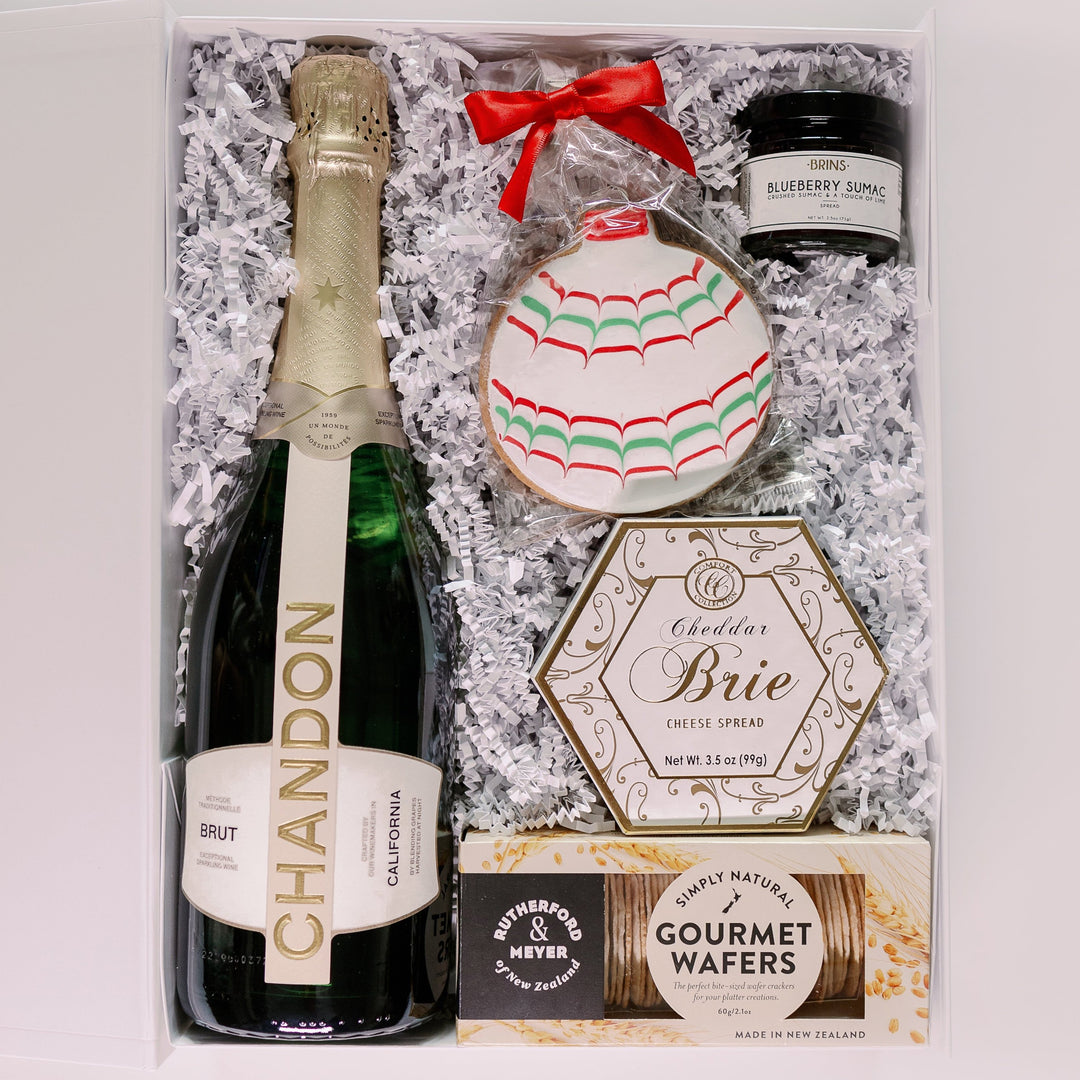 Studio photograph of a luxury holiday gift box featuring a bottle of Chandon Brut, a hexagonal box of Cheddar Brie, and a festive ornament-shaped cookie with red and green scalloped icing and a bright red ribbon bow nestled in white crinkle paper