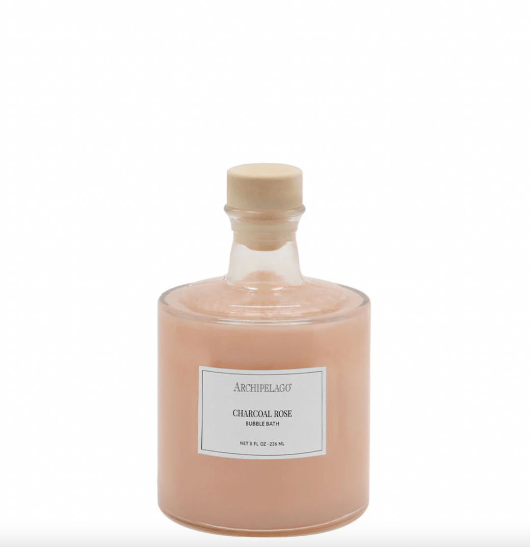 Product photograph by Archipelago featuring an apothecary glass bottle of Charcoal Rose Bubble Bath with a minimalist brand label and light wood stopper filled with peach liquid on a white backdrop