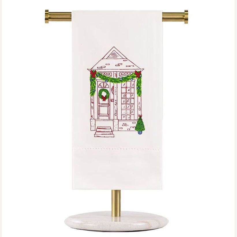 Embroidered linen hand towel by Hibiscus Linens featuring a festive boutique storefront entrance adorned with green garlands, a red-bowed wreath, and a small potted Christmas tree displayed on a gold and marble stand