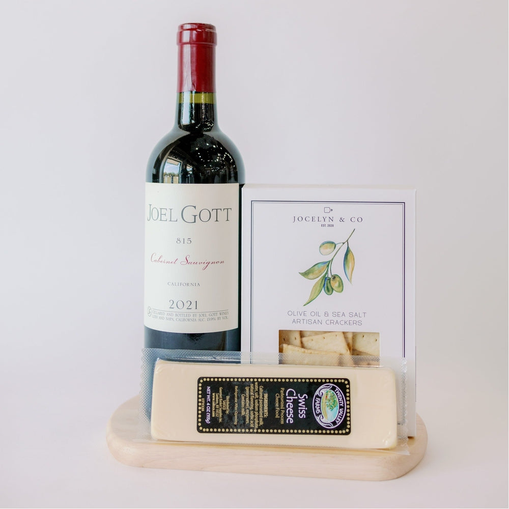 Digital photograph of a gourmet wine and cheese gift set featuring a bottle of Joel Gott 815 Cabernet Sauvignon, Jocelyn & Co artisan crackers, and Twenty Valley Farms swiss cheese on a round wooden board, highlighted by a crimson bottle seal and a green watercolor botanical illustration