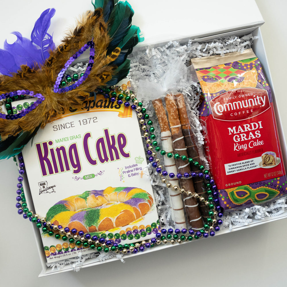 Photograph of a Louisiana Mardi Gras gift box featuring Mam Papaul's King Cake mix and Community Coffee embellished with a vibrant feathered masquerade mask and iconic purple, green, and gold beaded necklaces nesting in white shredded paper