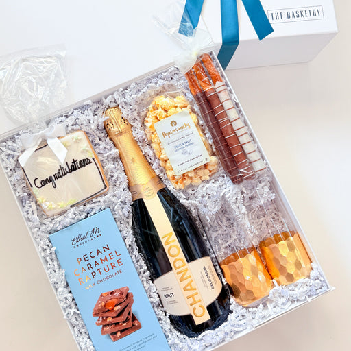 A luxury champagne gift hamper presented in a white box with a blue ribbon, filled with a bottle of champagne, a chocolate bar, a bag of pecan brittle, and two orange glasses on white crinkle paper.