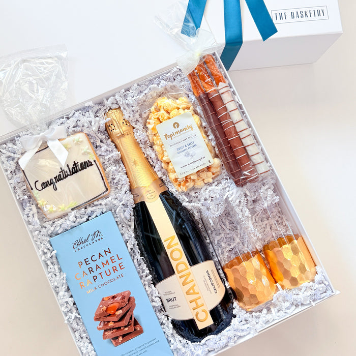A luxury champagne gift hamper presented in a white box with a blue ribbon, filled with a bottle of champagne, a chocolate bar, a bag of pecan brittle, and two orange glasses on white crinkle paper.