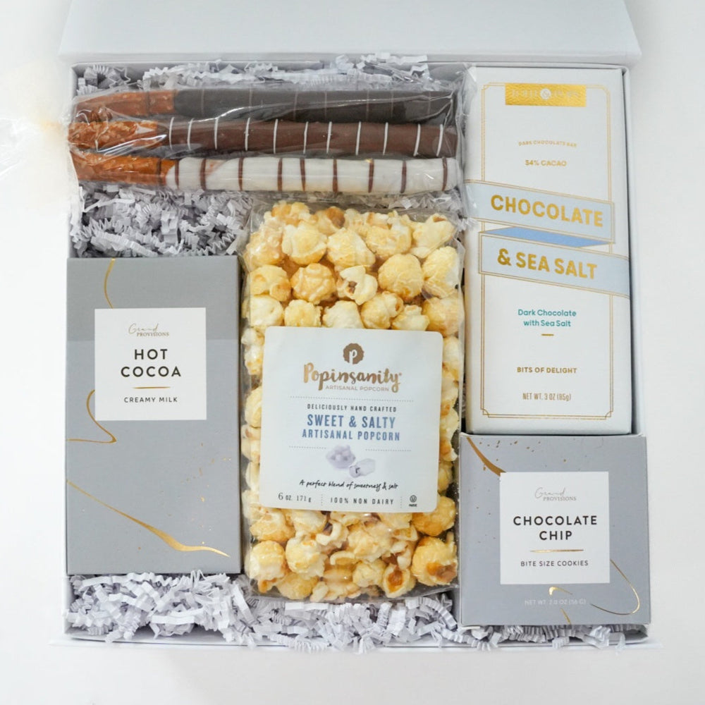 Product photograph of a Grand Provisions gourmet gift box featuring Popinsanity sweet and salty popcorn, three assorted chocolate-drizzled pretzel rods, and a chocolate and sea salt bar nestled in white crinkle paper alongside grey boxes with gold-foil accents