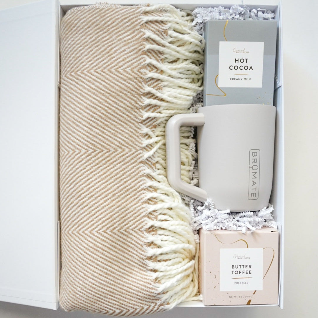 Gift set with beige blanket, mug, and hot cocoa mix in a box on a light background