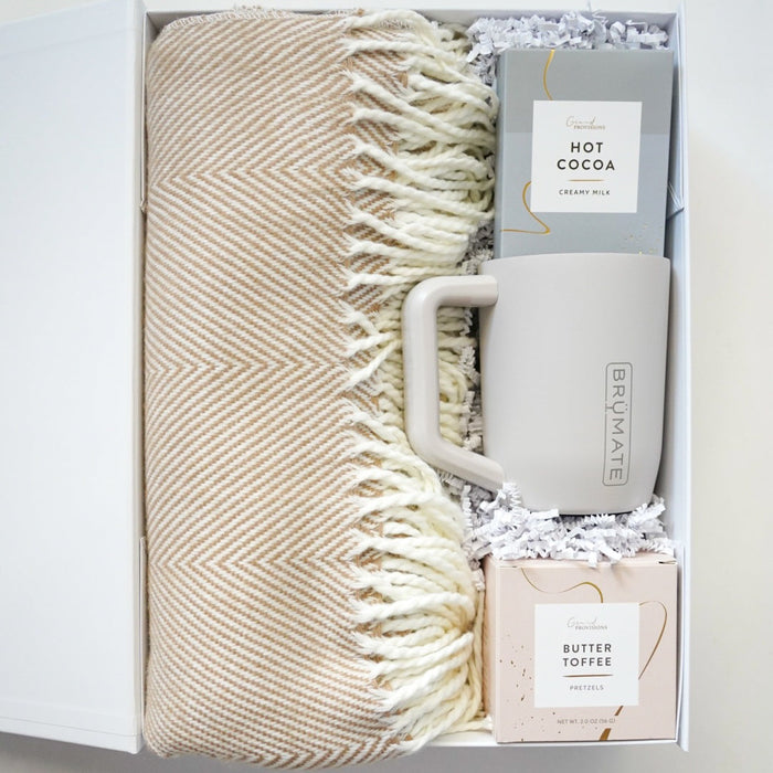 Gift set with beige blanket, mug, and hot cocoa mix in a box on a light background