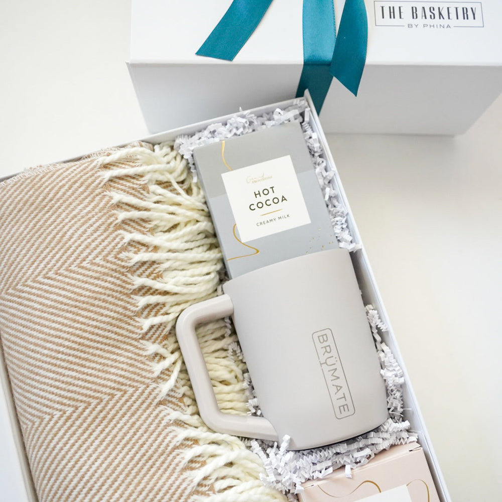 Digital photograph by The Basketry by Phina of a curated winter gift box featuring a tan herringbone-patterned throw blanket, a BrüMate insulated mug, and creamy milk hot cocoa mix accented with a teal blue ribbon