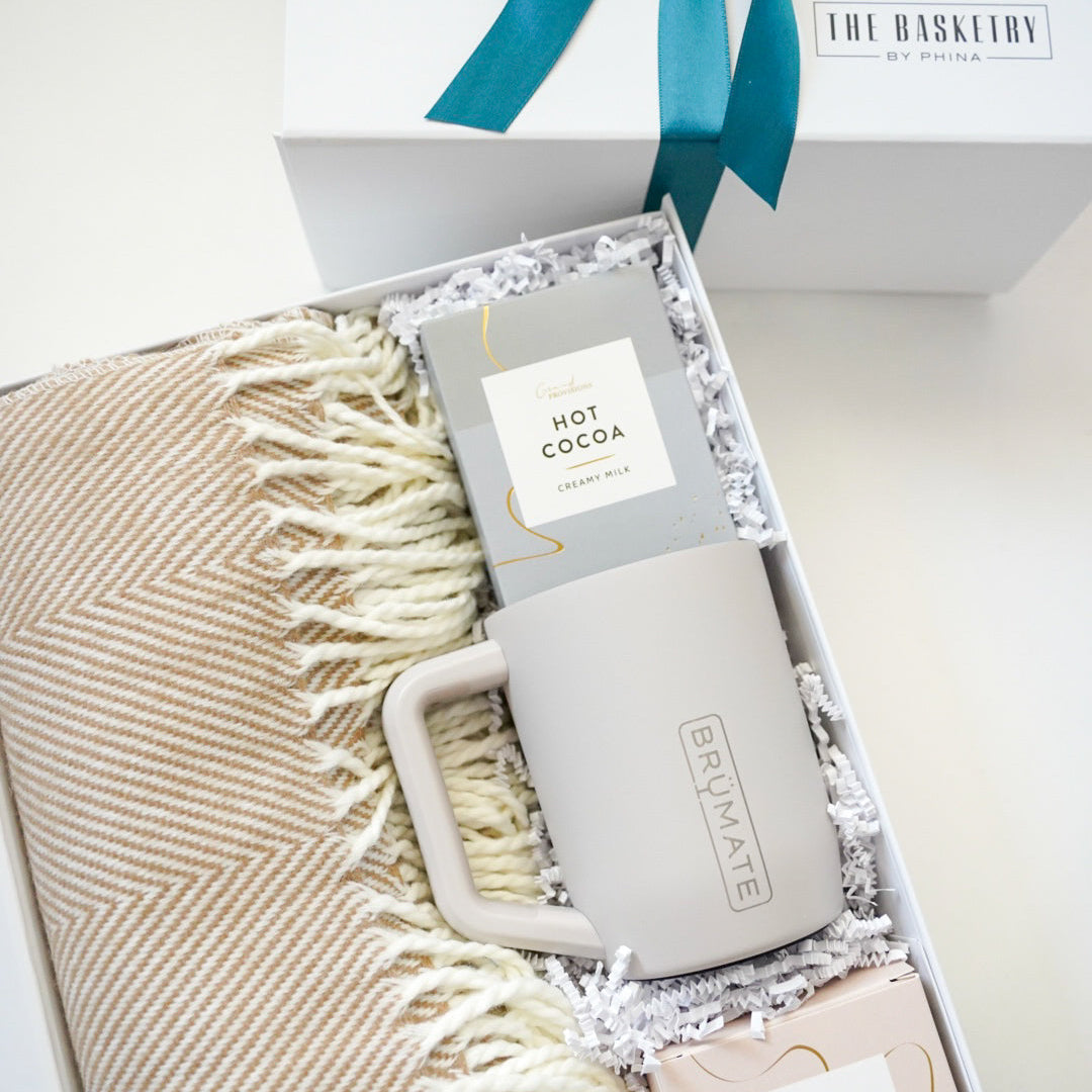 Digital photograph by The Basketry by Phina of a curated winter gift box featuring a tan herringbone-patterned throw blanket, a BrüMate insulated mug, and creamy milk hot cocoa mix accented with a teal blue ribbon