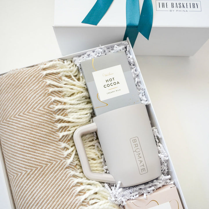 Digital photograph by The Basketry by Phina of a curated winter gift box featuring a tan herringbone-patterned throw blanket, a BrüMate insulated mug, and creamy milk hot cocoa mix accented with a teal blue ribbon