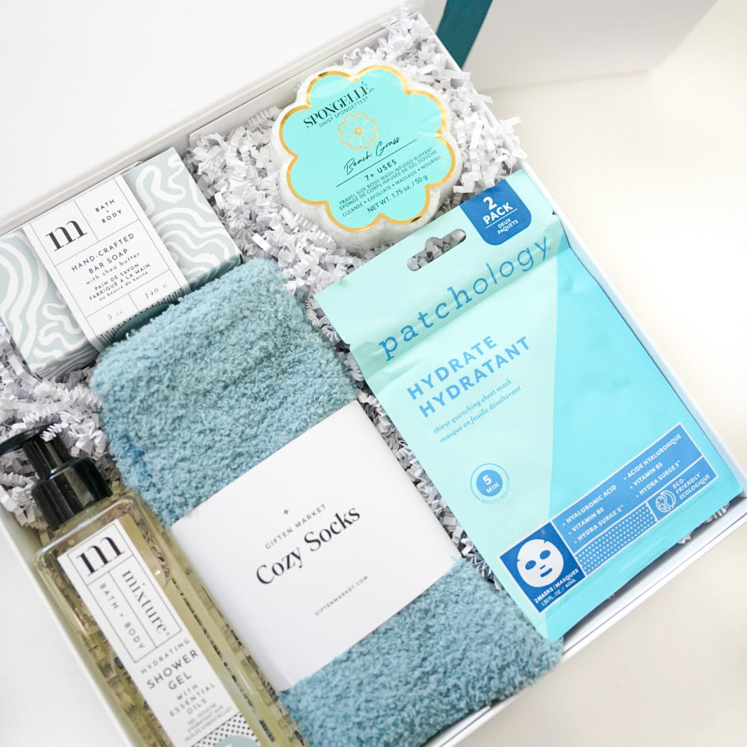 Digital product photograph of a Giften Market self-care gift box featuring Patchology hydrate masks, a Spongellé beach grass buffer, and Mixture bath products accented by soft light blue fuzzy socks and white crinkle paper filler