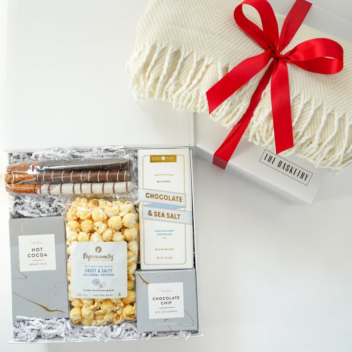 Product photograph by The Basketry of a gourmet gift box on a white surface featuring Popinsanity popcorn and chocolate-covered pretzels alongside a cream-colored fringed blanket adorned with a vibrant red satin bow