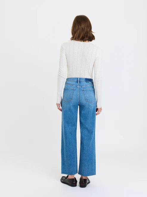 Photograph of a woman standing in a bright white studio, featuring high-waisted blue wide-leg jeans with a dark blue waistband patch, a white long-sleeved pointelle knit top, and dark brown leather loafers